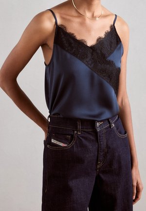 Persoon draagt een marineblauwe satijnen top met zwarte kanten afwerking en donkerblauwe high-waisted jeans, aangevuld met een dunne gouden ketting.