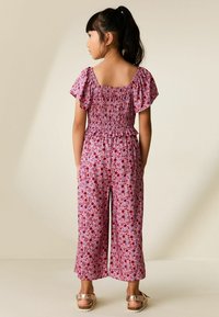 Sötét hajú lány lófarokban, rózsaszín virágmintás ráncolt felsőt és hozzá illő bő szárú nadrágot visel, háttal áll, kezét a zsebében tartva.