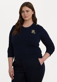 Marineblå kabelstrikket genser med ribbestrikket nederkant og mansjetter. Har gulllogo detaljer på venstre side og dekorative skulderknapper.