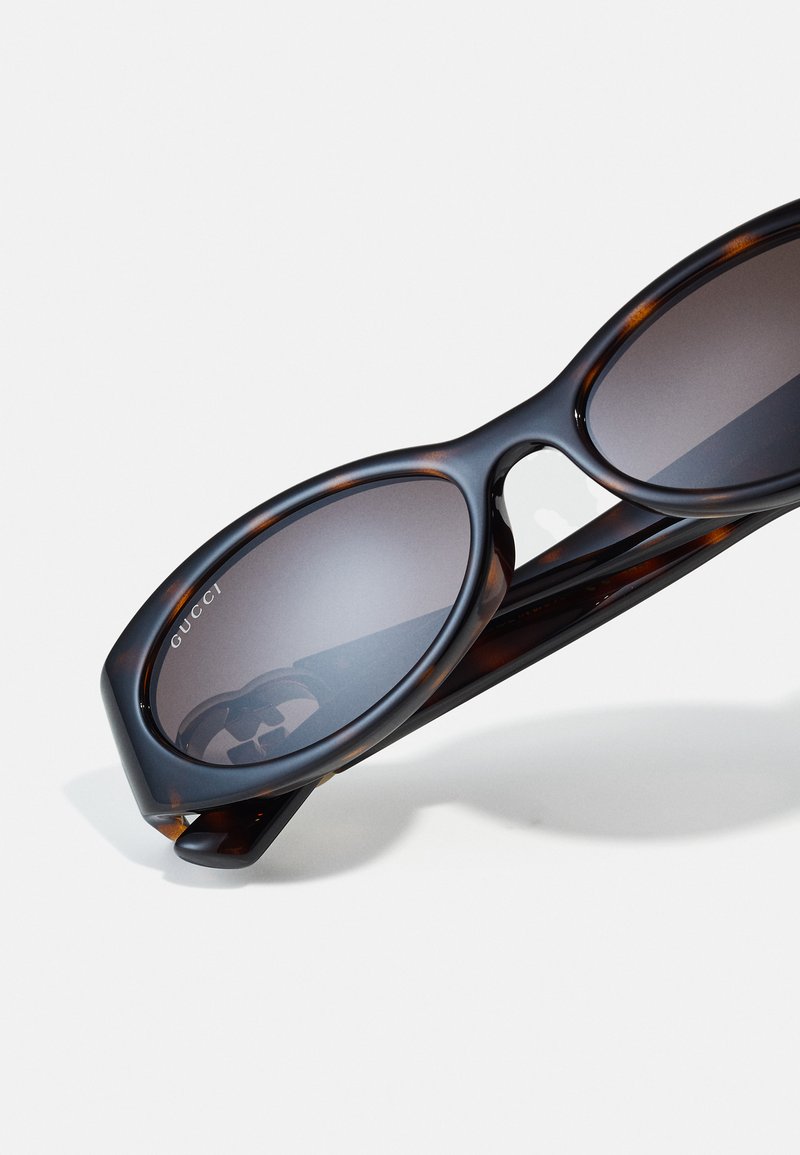 Gucci sunglasses featuring a tortoiseshell pattern, oval shape, grey lenses, and smooth plastic frames. Logo displayed on the lens.