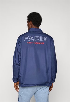 Man wearing navy blue jacket with "PARIS SAINT-GERMAIN" text on back, paired with light gray cargo pants, standing against plain background.