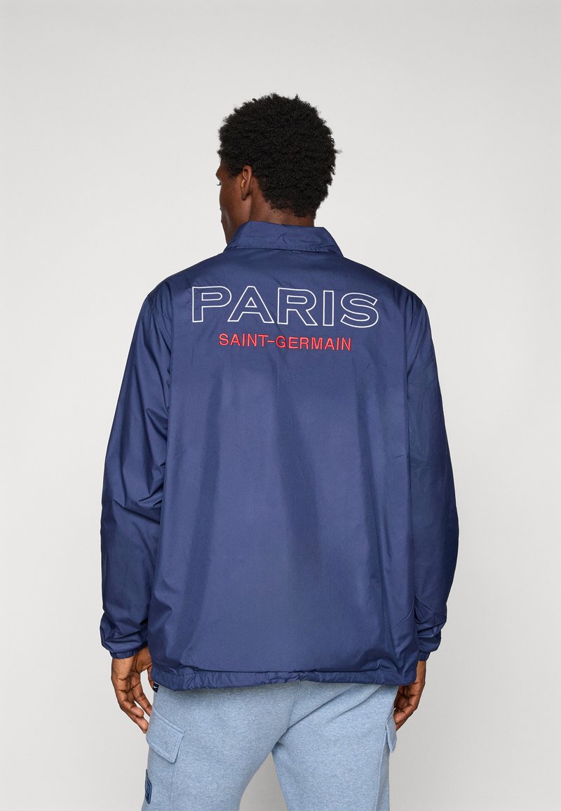Man wearing navy blue jacket with "PARIS SAINT-GERMAIN" text on back, paired with light gray cargo pants, standing against plain background.