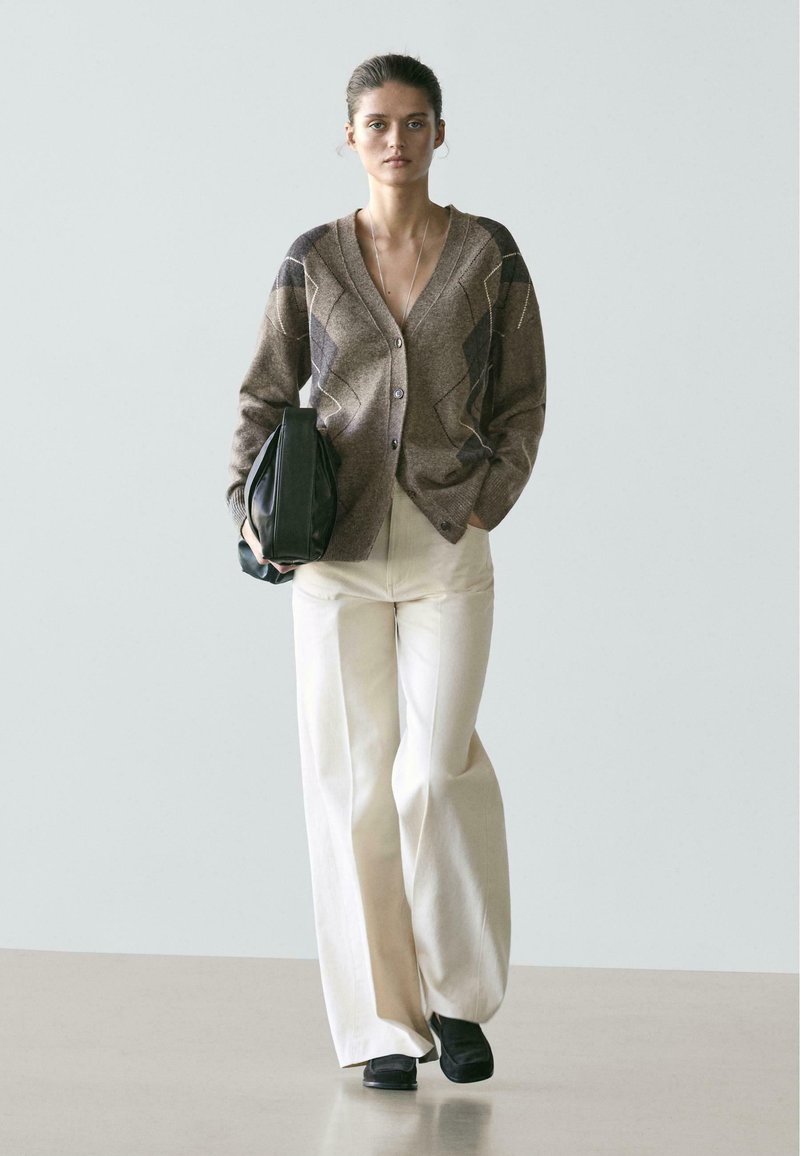 Kobieta idąca, ubrana w brązowy kardigan w wzory, białe szerokie spodnie i czarne buty, trzymająca dużą czarną torbę, na tle jasnego, jednolitego tła.