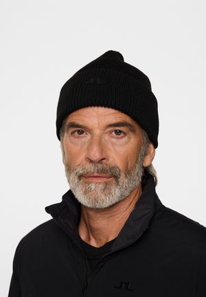 Bonnet tricoté noir avec une texture côtelée et un sommet pointu, arborant un logo discret. Porté avec une veste noire zippée.