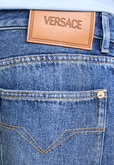 Blue denim jeans with a stitched back pocket and a tan leather label embossed with "VERSACE" above the pocket.