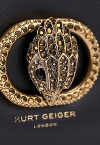Accessorio in pelle nera con chiusura in tonalità oro, arricchita da embellishments sfaccettati intricati e un design dettagliato e testurizzato. Logo "Kurt Geiger" visibile.