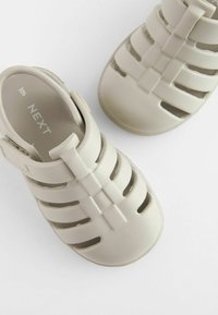 Helle beige Kindersandalen aus strukturiertem Gummi, offene Zehenpartie, mehrere Belüftungsschlitze und ein Klettverschluss zur Befestigung.