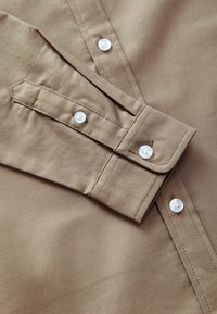 Beige koše�ľa s golierom na gombíky, manžetami a bielymi plastovými gombíkmi. Materiál má hladký, štruktúrovaný povrch s viditeľným stehom.