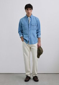 Camisa de mezclilla azul claro con botones en la parte delantera y dos bolsillos en el pecho, combinada con pantalones color marfil y zapatos marrón oscuro. Sosteniendo una gorra marrón.