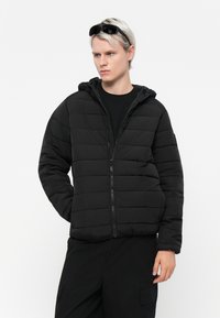 Veste matelassée noire avec une fermeture éclair sur le devant, des coutures horizontales et une capuche. Le modèle porte un t-shirt noir et des lunettes de soleil. Texture mate.