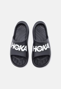 Ζευγάρι μαύρα σανδάλια Hoka slip-on με υφασμένα πέλματα και λευκό λογότυπο στα λουράκια, εκτεθειμένα σε λευκό φόντο.