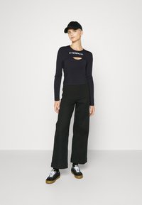 Musthave musta pikkade varrukatega crop top, millel on ees lõikeauk ja valge "ETERNAL" tekst, koos laienevate mustade pükste ja mustade kummist talladega tossudega.