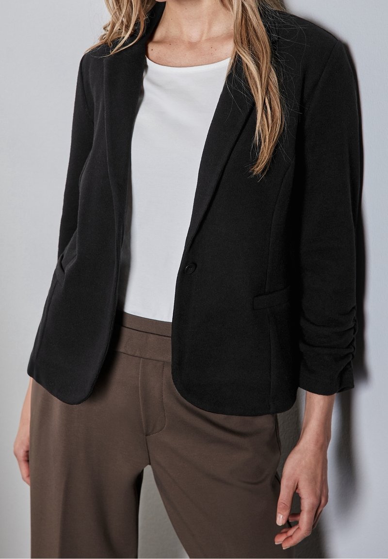 Svart kavaj med skräddarsydd passform, långa ärmar, en knappstängning och sidofickor. Bärs över en vit topp och bruna byxor.