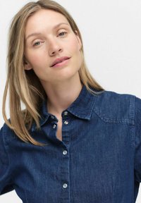 Mörkblå denimskjorta med spetsig krage, silvriga knappar och en mjuk textur. Har förstärkningsstygn på axlarna.