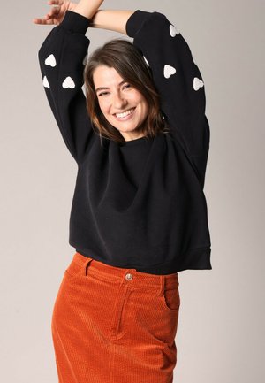 Sweatshirt noir court avec des motifs de cœurs blancs sur les manches, associé à une jupe en velours côtelé orange avec une fermeture à boutons sur le devant.