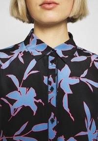 Una vista ravvicinata di una persona che indossa una camicia con colletto caratterizzata da uno sfondo nero con audaci motivi floreali blu e rosa.