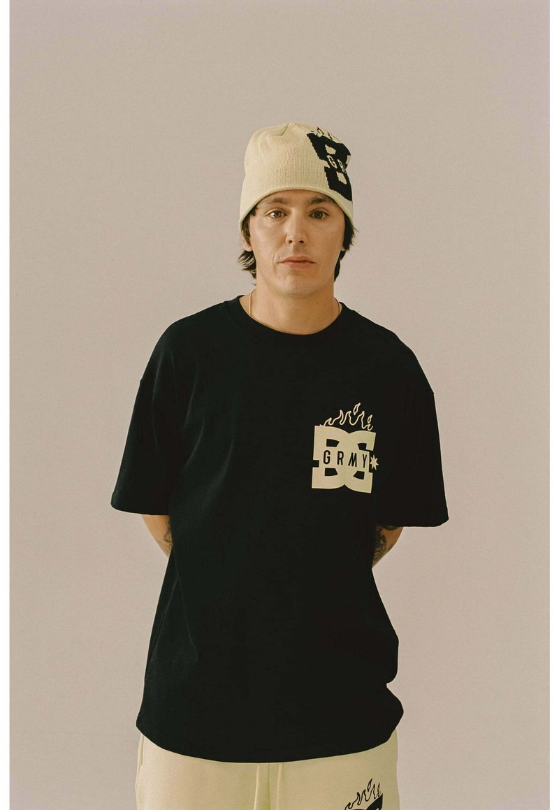 Jongeman in beige muts en zwart T-shirt met "GRMY" logo, staand tegen een effen lichte achtergrond met de handen achter de rug.