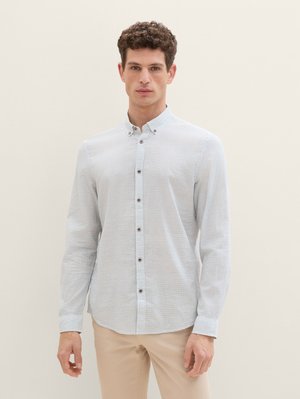 Lichtblauw overhemd met een subtiele streepjespatroon, lange mouwen en bruine knopen, gedragen met beige broek.
