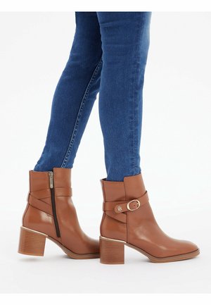 Personne portant un jean skinny bleu et des bottines en cuir marron à talons blocs avec fermeture éclair latérale sur fond blanc.