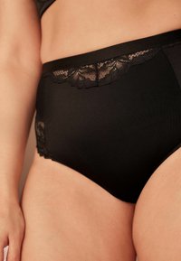 Must have kõrge talje mustad aluspüksid, mis on valmistatud siledast kangast, ja millel on lisadetaile ja tekstuuri andev pitsääris vööl ja külgedel.