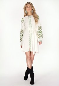 Valkoinen mekko, jossa on vihreää geometristä brodeerausta hihoissa ja edessä, nyörikaulus, kapea vyötärö ja reiden ylle ulottuva halkio. Mustat nilkkurit.