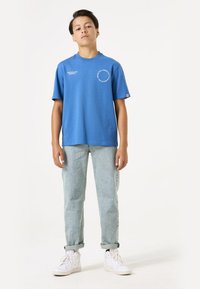 Camiseta de algodón azul con mangas cortas, cuello redondo y detalle de texto en la parte frontal. Combinada con Jeans de mezclilla de lavado claro y zapatillas blancas.