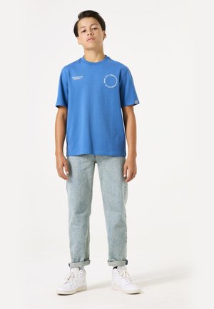 Camiseta de algodón azul con mangas cortas, cuello redondo y detalle de texto en la parte frontal. Combinada con Jeans de mezclilla de lavado claro y zapatillas blancas.