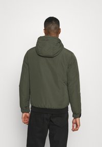 Giacca imbottita verde scuro con cappuccio, polsini ed elastico in vita. Texture esterna liscia con finitura opaca, senza motivi visibili.