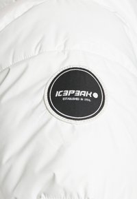 Vit vadderad jacka med en svart rund lapp fram som har texten "ICEPEAK" och "ESTABLISHED IN 1996" sydd på. Slät yta.