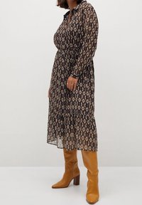 Vestido largo y transparente con un diseño estampado en tonos oscuros. Presenta un cuello, mangas largas, una cintura definida y una falda en capas. Combinado con botas marrones hasta la rodilla.