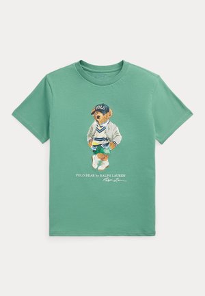 Grøn kortærmet t-shirt med en grafik af en bjørn iført kasket, jakke, sweater, shorts og sneakers, med tekst under bjørnen.