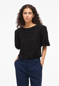 Melna teksturēta blūze ar krokainu dizainu, īsām piedurknēm un apaļu kakla izgriezumu, apvienota ar tumši ziliem biksēm.