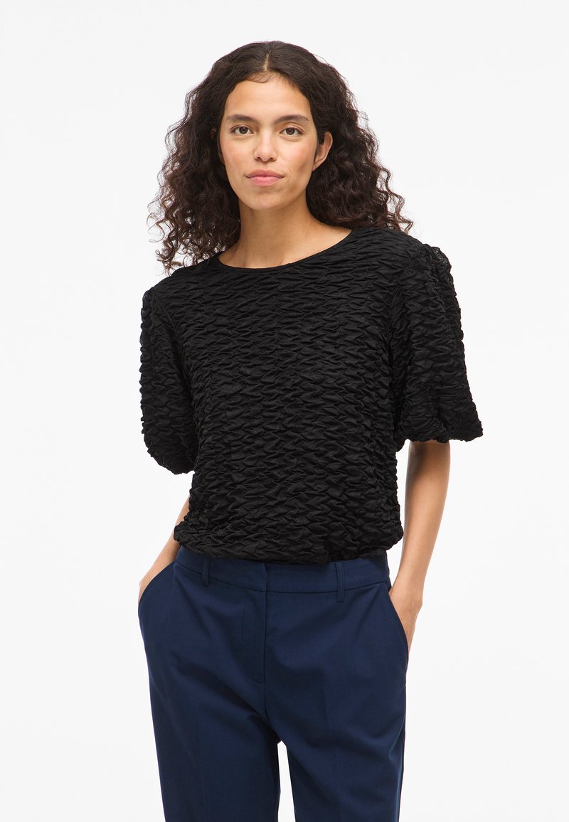 Melna teksturēta blūze ar krokainu dizainu, īsām piedurknēm un apaļu kakla izgriezumu, apvienota ar tumši ziliem biksēm.