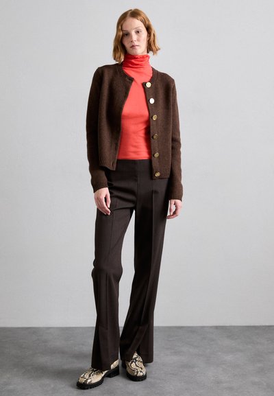 Brown cardigan with gold buttons over a coral turtleneck, paired with brown tailored pants and snake-print shoes, on a light gray backdrop.