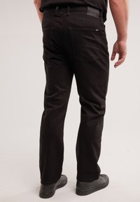 Férfi fekete egyenes szárú farmernadrágban és fekete cipőben, hátulnézetből, egyszínű világos háttér előtt.