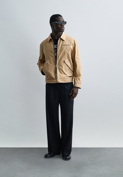 Άνδρας που φοράει μπεζ μπουφάν, μαύρο μπλουζάκι με σχέδιο, φαρδιά μαύρη παντελόνια, μαύρα παπούτσια και γυαλιά ηλίου, στέκεται μπροστά σε απλό γκρι φόντο.