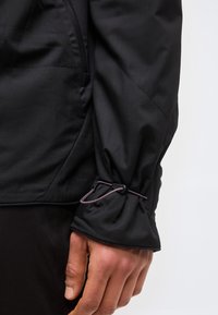 Prim-plan al unei mâini care ajustează mâneca unei jachete negre cu un șnur elastic și un dispozitiv de fixare pe un fundal deschis la culoare.