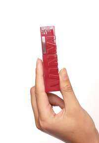 Maybelline Superstay Vinyl Ink i djärv röd förpackning, rektangulär form, glansig yta, prägling i bokstäverna, hålls i en hand mot en vit bakgrund.