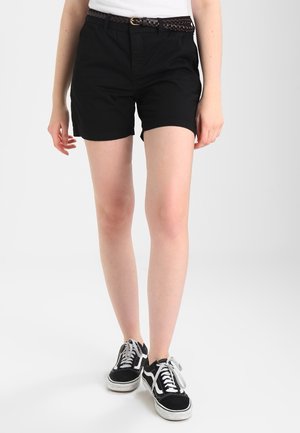 Person iført sorte shorts med flettet bælte og sort-hvide sneakers, der står mod en ensfarvet lys baggrund.