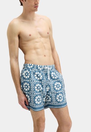 Skjorteløs mand iført blå og hvide mønstrede badeshorts med snøre, stående mod en ensfarvet lys baggrund.