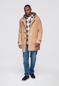 Sandfärgad duffelrock med träknäpplås, två framfickor och foder. Bärs över en rutig skjorta, kombinerad med blå jeans och mörka skor.