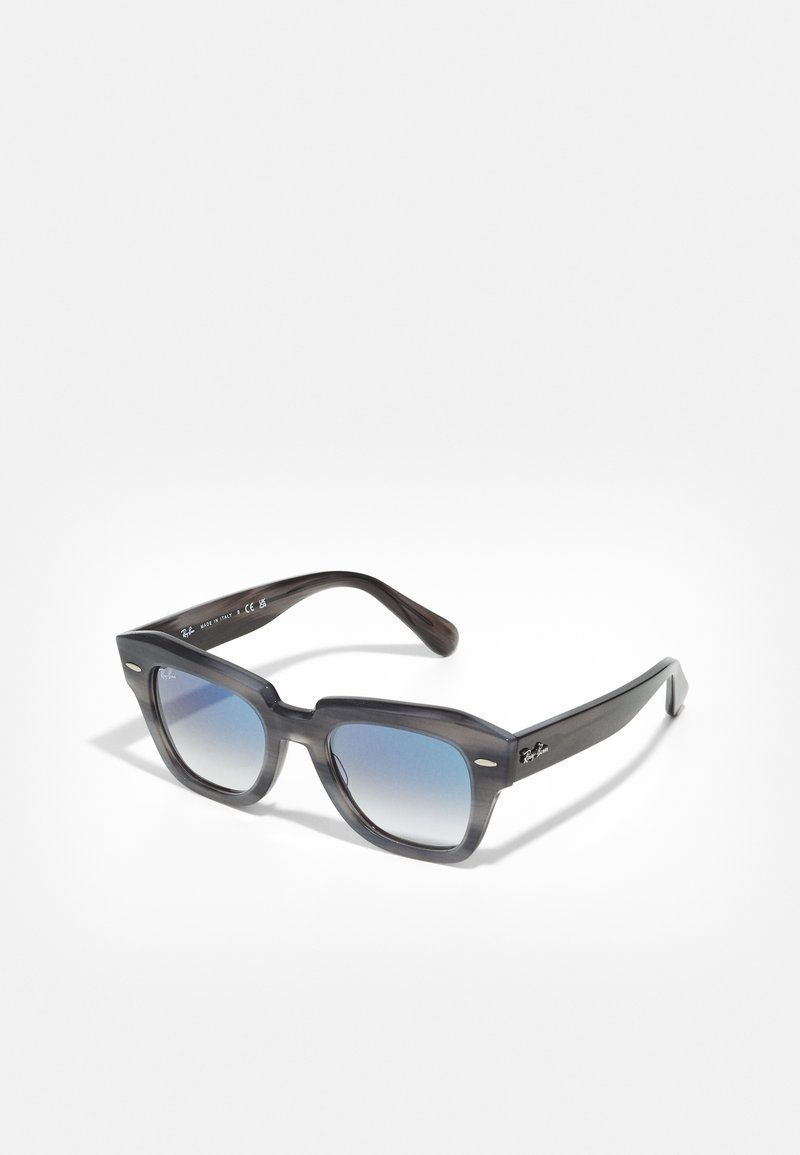 Ochelari de soare pătrați cu ramă în gradient gri și lentile în gradient albastru. Caracteristici includ accente metalice și branding subtil pe brațe.