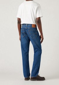 Jeans de mezclilla azul con un corte recto, bolsillos traseros visibles y una etiqueta de cuero en la cintura. Combinados con una camiseta blanca de manga corta.