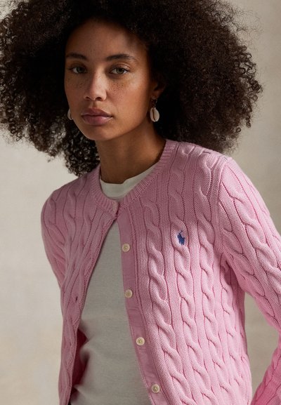 Moteris su garbanotais plaukais, dėvinti auskarus su kriauklėmis ir rožinį megztinį su pintinėmis raštais ant baltos palaidinės, prieš neutralią foną.