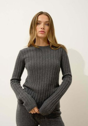 Pull en maille côtelée gris avec des manches longues et ajustées. Texture douce et encolure ronde. Le modèle tient l'ourlet en posant.