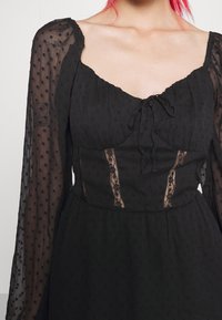 Black dress with textured fabric, sheer long sleeves, lace accents, and a square neckline. Features a tie detail at the front.