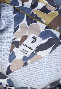 Kleurrijke stof met patronen en abstracte vormen in marineblauw, beige en olijfgroen, met een label waarop de tekst "J&Joy, Ontworpen in België" staat.