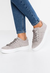 Piedi in sneakers in suede grigio chiaro con decorazioni di perle e lacci in satin, abbinati a jeans slim-fit blu su uno sfondo bianco.