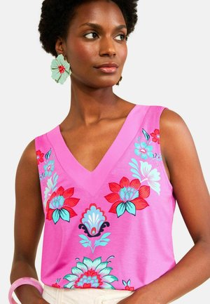 Kobieta w różowym topie bez rękawów z czerwonymi, niebieskimi i białymi kwiatowymi wzorami, dużymi niebiesko-czerwonymi kolczykami w kształcie kwiatów i różową bransoletką.