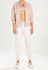 Giacca bomber zip-up rosa chiaro con collo rotondo, abbinata a un reggiseno sportivo beige, pantaloni della tuta bianchi e scarpe slip-on beige.